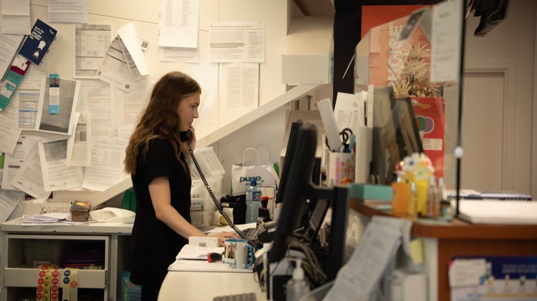 A pharmacy assistant at the back counter, working between a screen and a wall covered in printed notes and prescriptions.