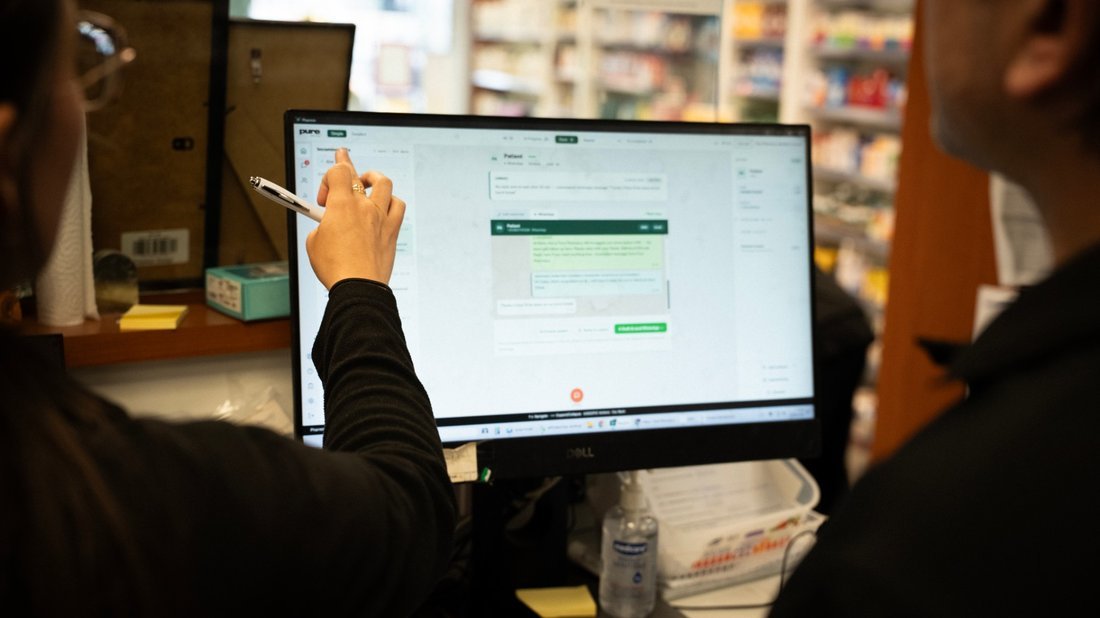A pharmacist pointing a pen at a message thread on the dispensary screen, a colleague at their shoulder.