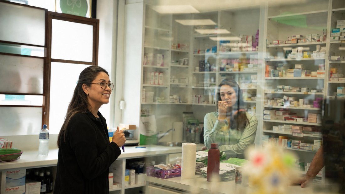 A pharmacist seen through the dispensary glass at the end of the day, talking to a colleague.